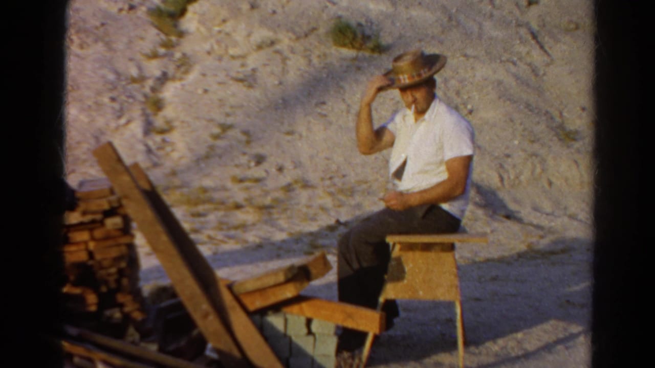 Man in a straw hat sits on a wooden stool beside stacked firewood and lumber.