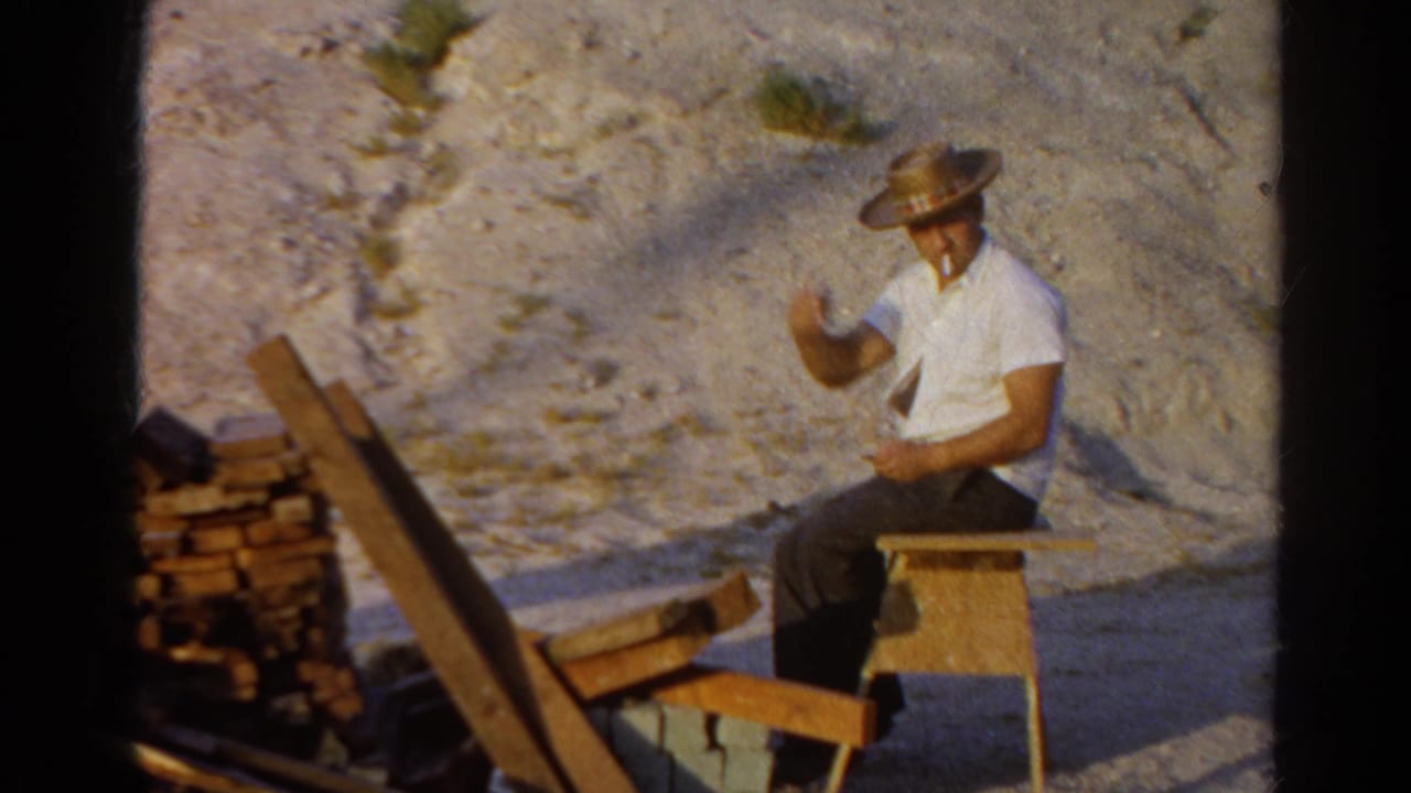 The man in the straw hat raises a hand toward the camera while seated at the outdoor Silver Lake work area.
