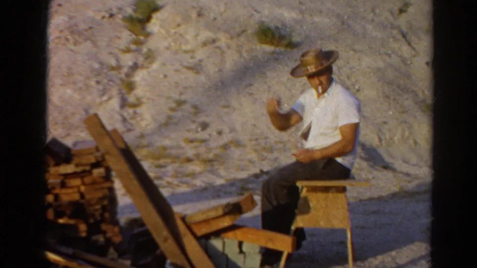 The man in the straw hat raises a hand toward the camera while seated at the outdoor Silver Lake work area.