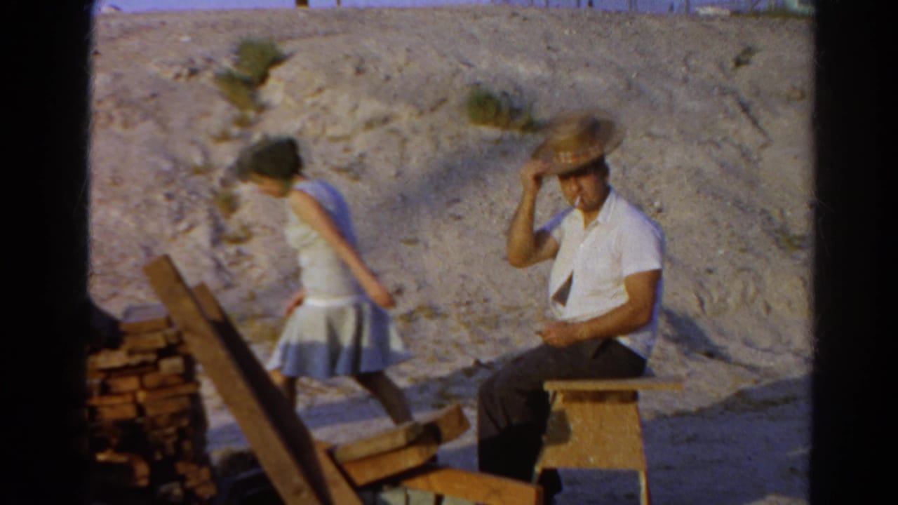 1966 Silver Lake home-movie frame showing a seated man in a straw hat and a woman walking away beside stacked wood in bright sun.