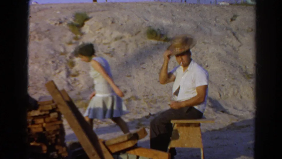 1966 Silver Lake home-movie frame showing a seated man in a straw hat and a woman walking away beside stacked wood in bright sun.