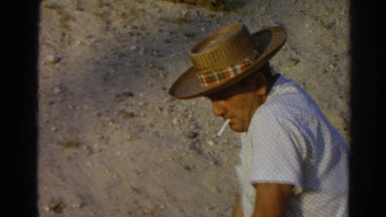 Closer view of a man in a straw hat and white shirt holding a cigarette against a dusty hillside.