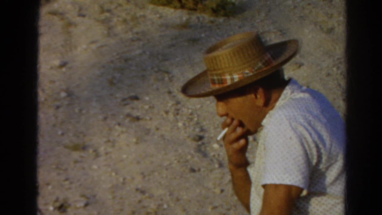 1966 Silver Lake home-movie frame showing the seated man touching a cigarette to his mouth in bright outdoor light.