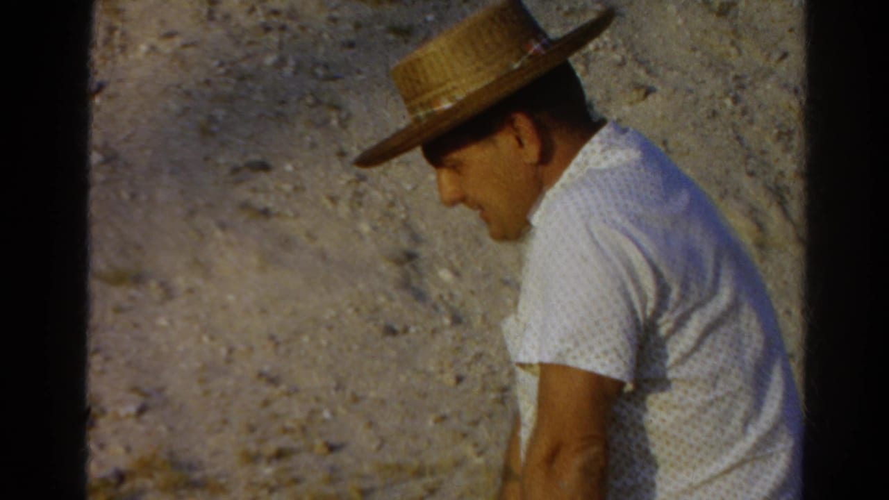 Seated man in straw hat and white shirt is shown in profile against gravel.