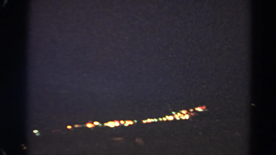 Soft, grainy 8mm frame showing a line of distant lights in a dark outdoor setting.