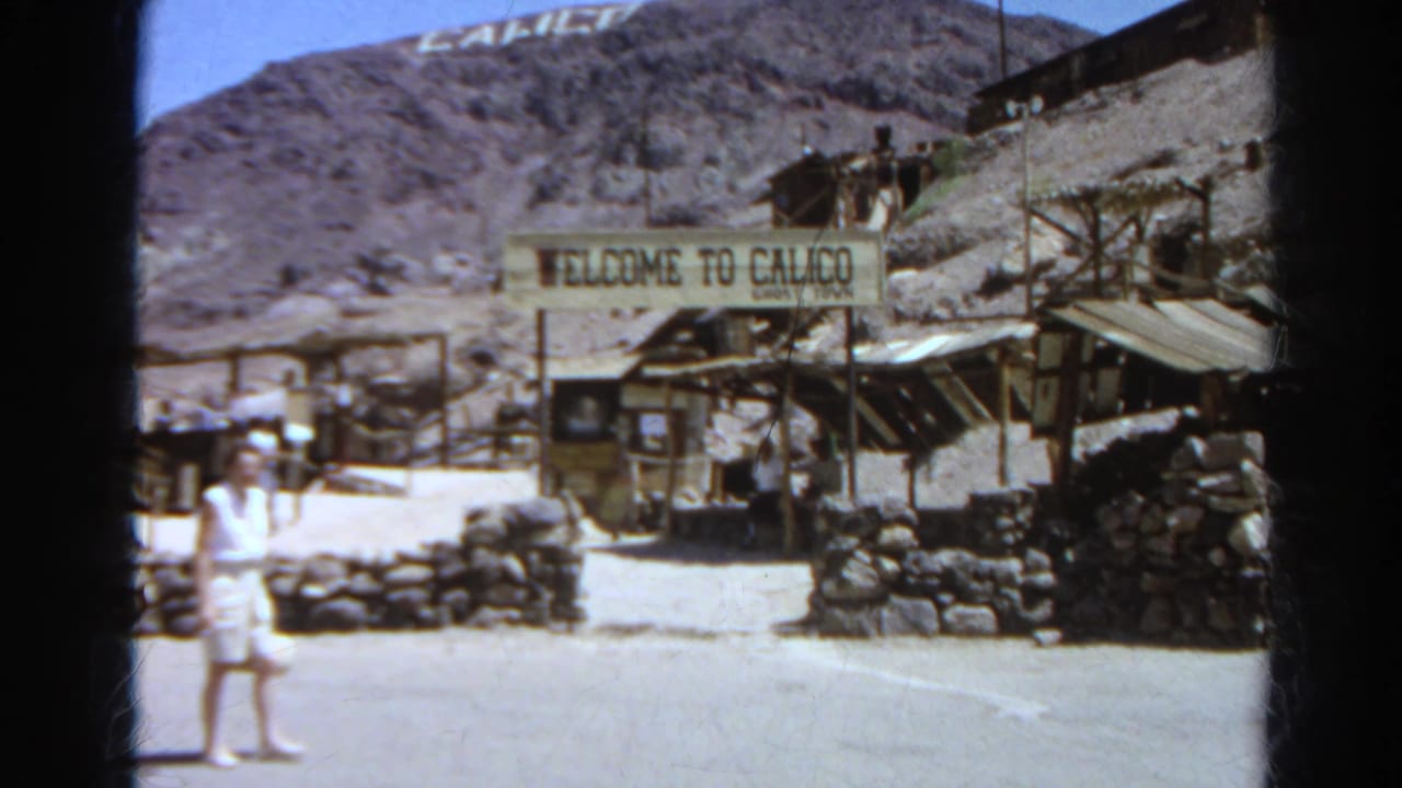 1966 home-movie frame of a woman in white near the Welcome to Calico Ghost Town sign, with arid hills and rustic structures behind.