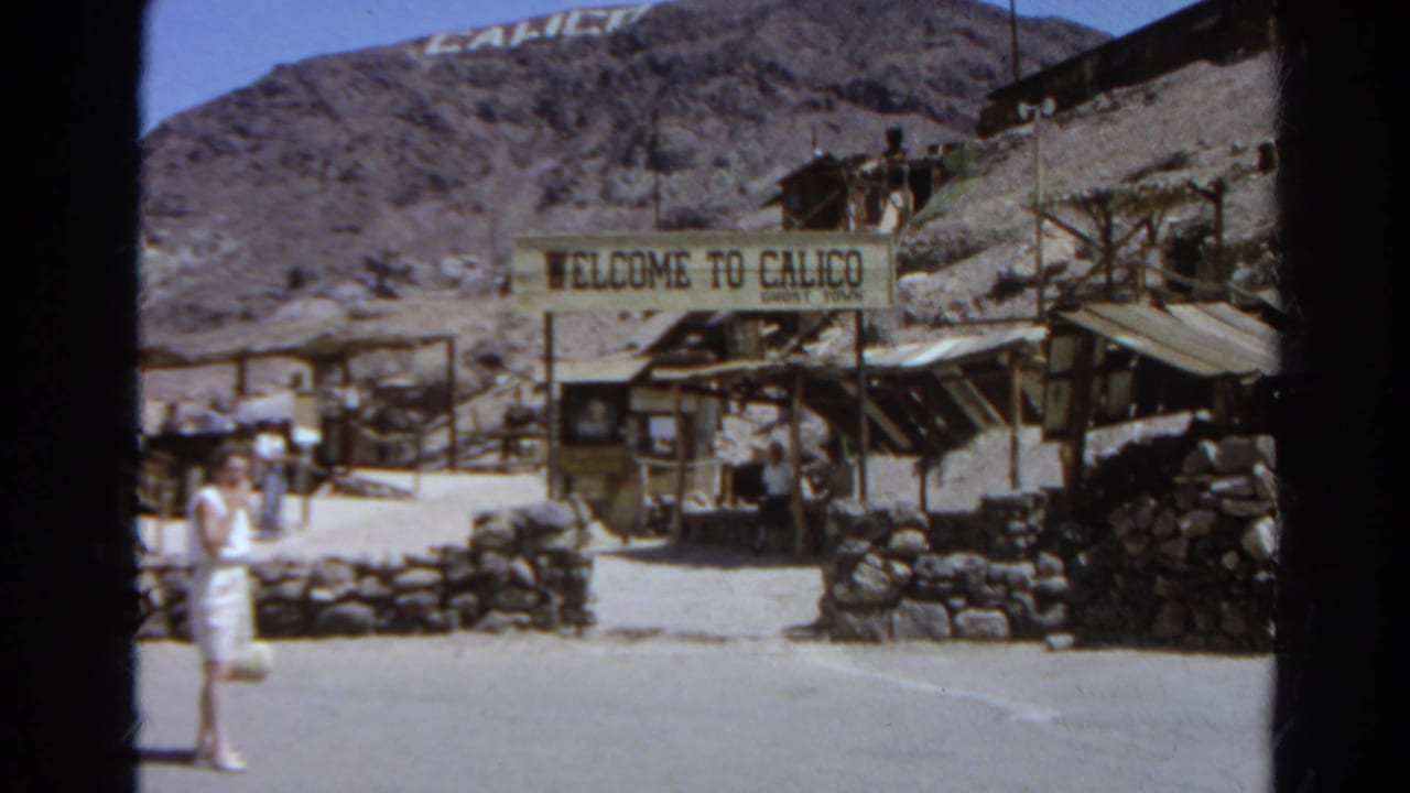 1966 8mm frame of the Calico Ghost Town entrance sign, stone walls, weathered buildings, and a woman partly visible at left.
