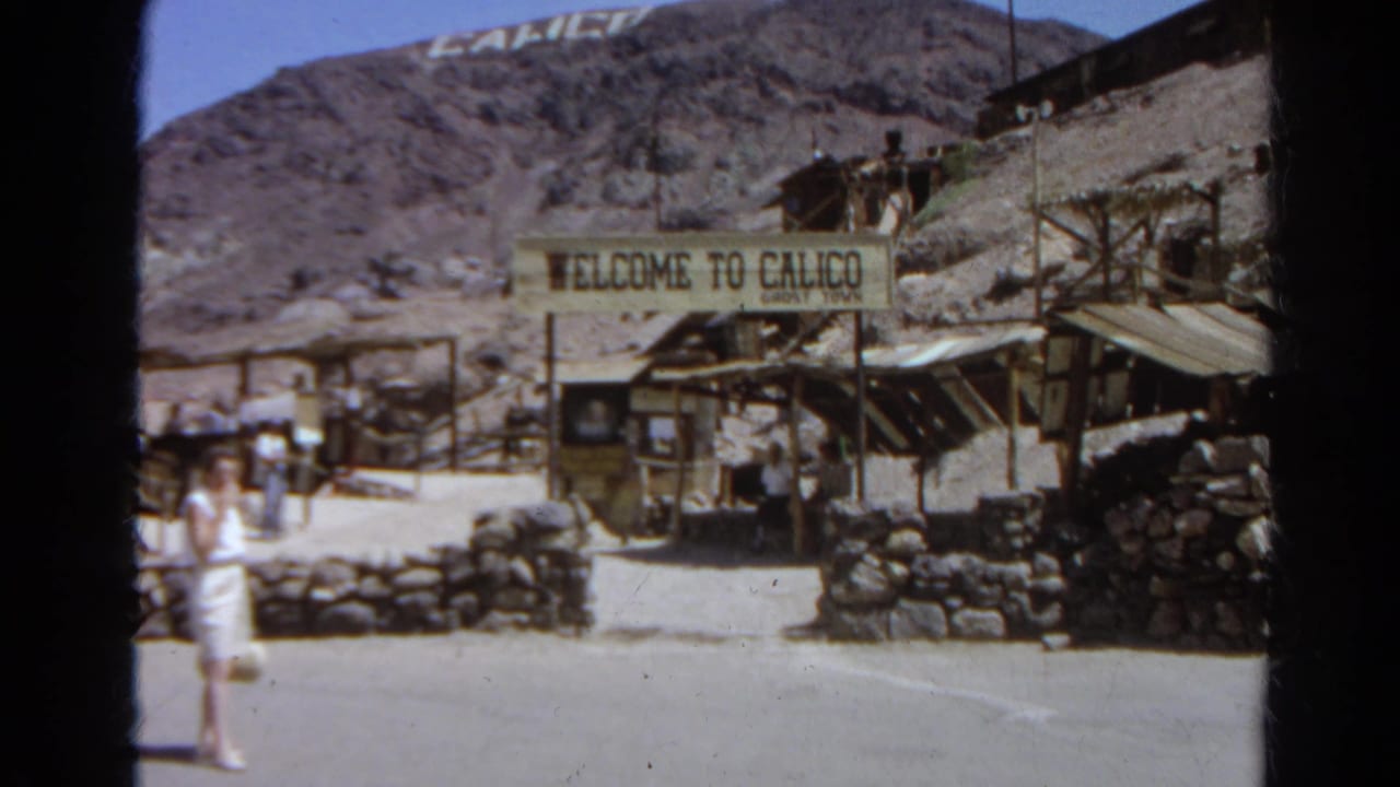 1966 8mm frame of tourists outside the Welcome to Calico Ghost Town entrance, with stone walls, wood structures, and desert hills visible.