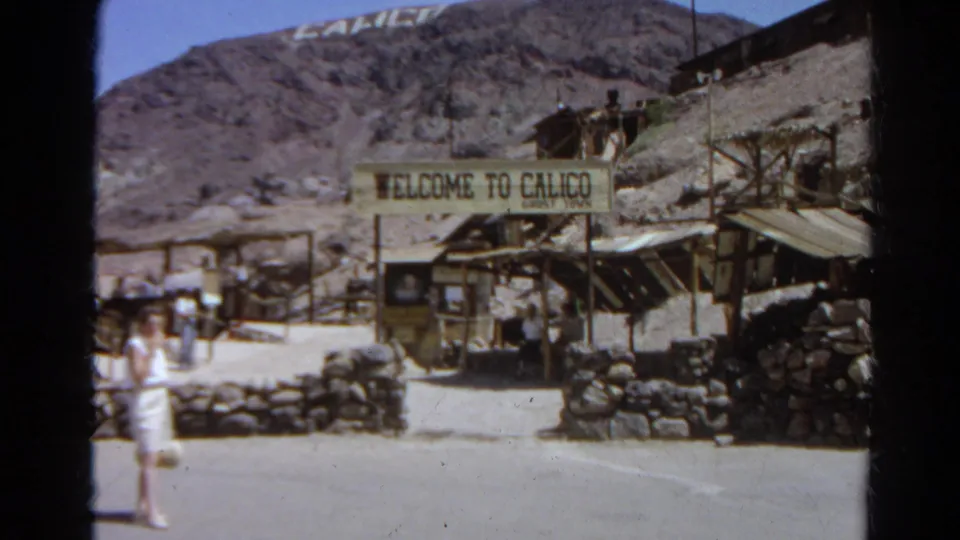 1966 8mm frame of tourists outside the Welcome to Calico Ghost Town entrance, with stone walls, wood structures, and desert hills visible.