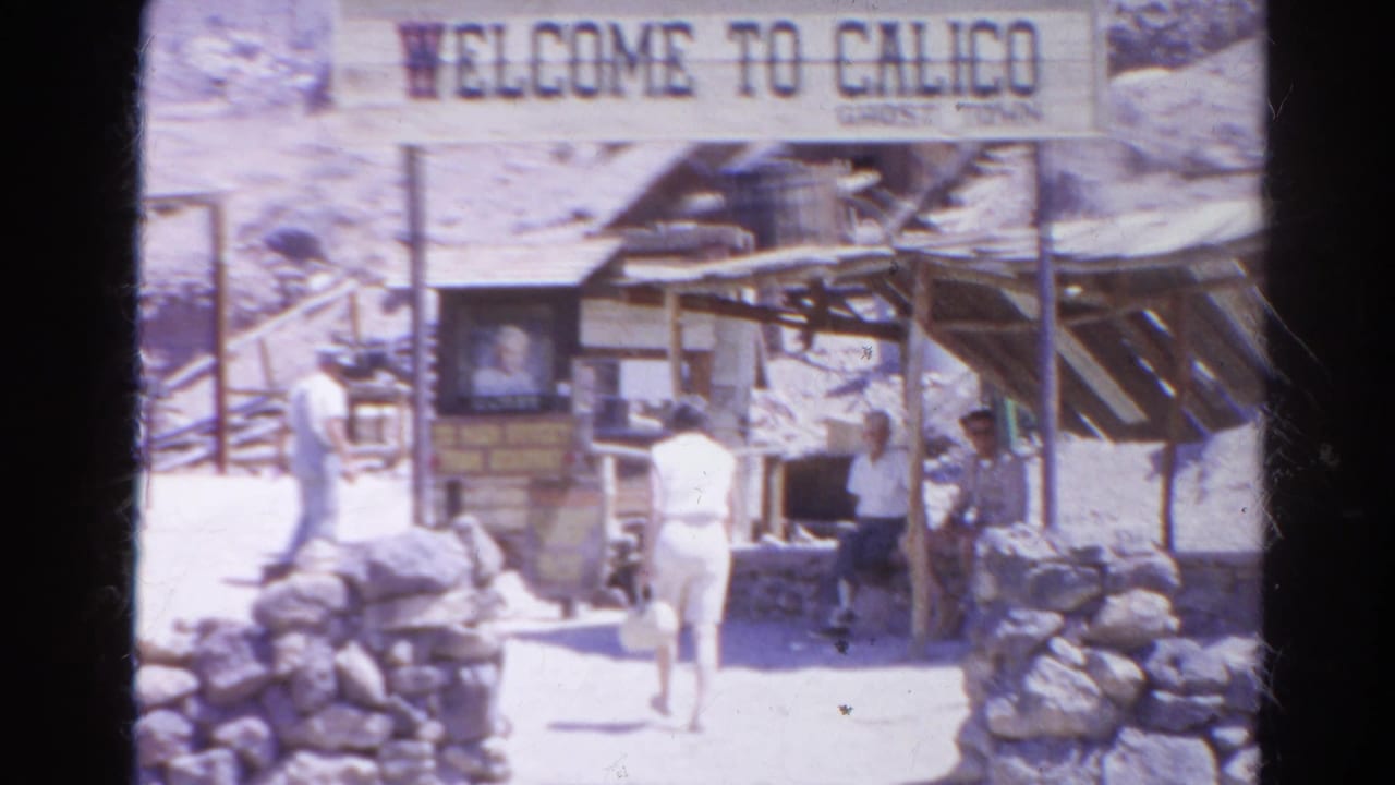 Tourists stand and walk beneath the Welcome to Calico Ghost Town sign near stone walls and a wooden awning.