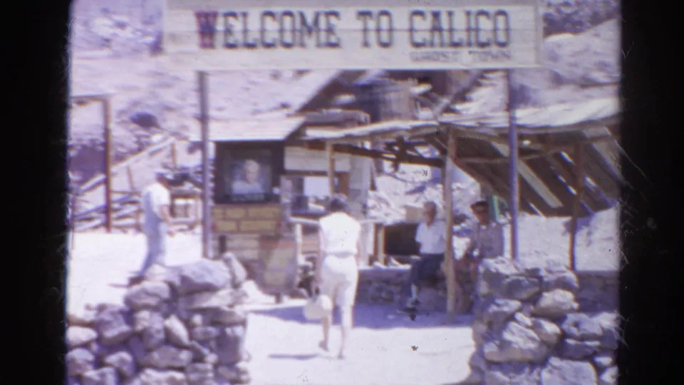 Visitors in 1960s clothing move through the Calico Ghost Town entrance area, with a readable welcome sign and rustic buildings in bright sun.