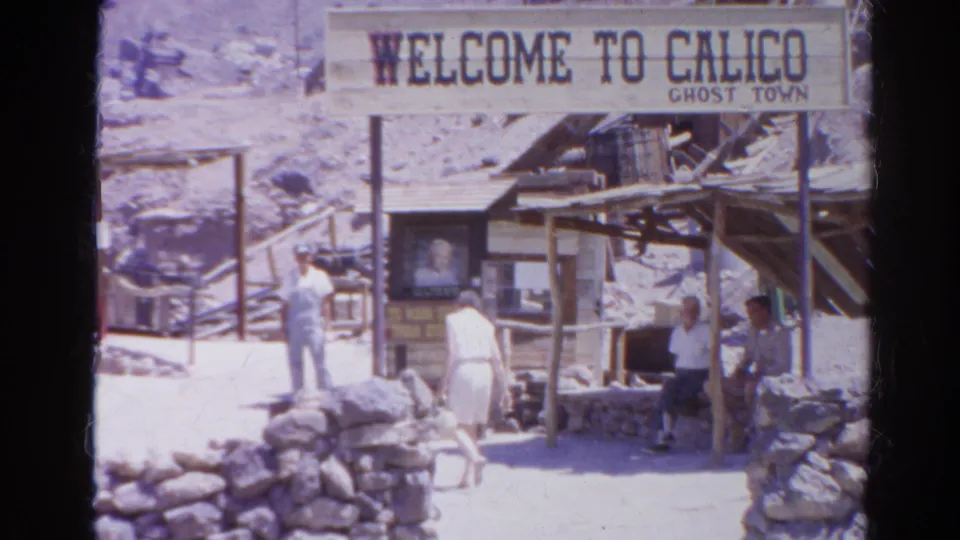 A closer 1966 home-movie view shows the Calico Ghost Town welcome sign, tourists, stonework, and timber structures in a desert setting.