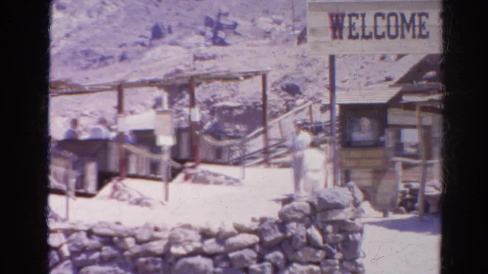 1966 Silver Lake, California home-movie frame showing visitors by open carts, a track area, stone wall, and partial Welcome sign.