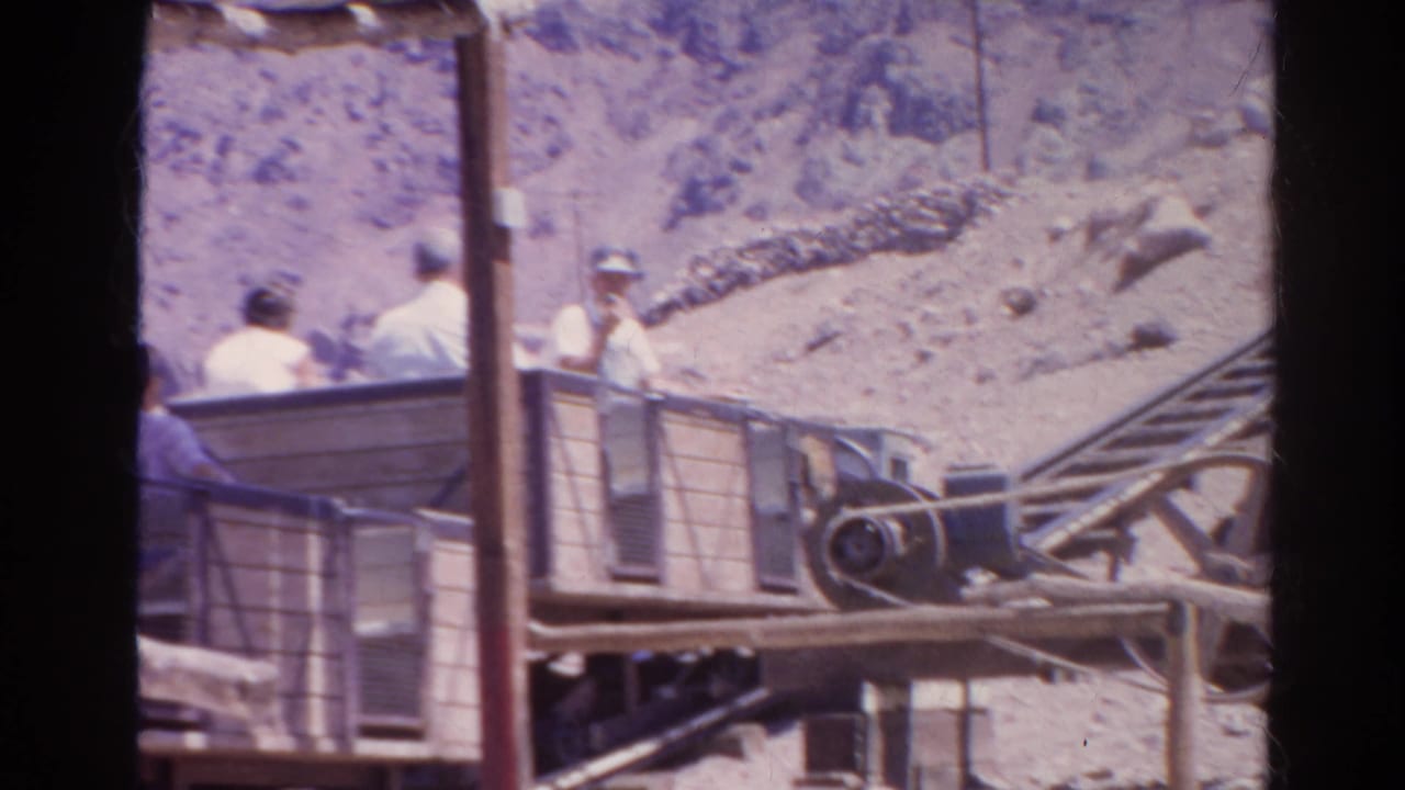 1966 Silver Lake, California home-movie frame showing riders in an open cart near rails, pulley hardware, posts, and rocky ground.