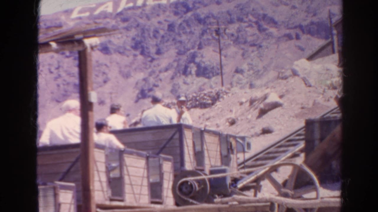A wooden cart with seated riders moves along a hillside track near pulley machinery.