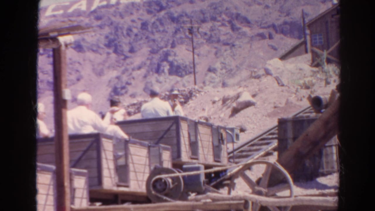 A home-movie frame shows the hillside cart lower in the shot, with riders, a large wheel, a partial sign, and a small wooden structure at right.