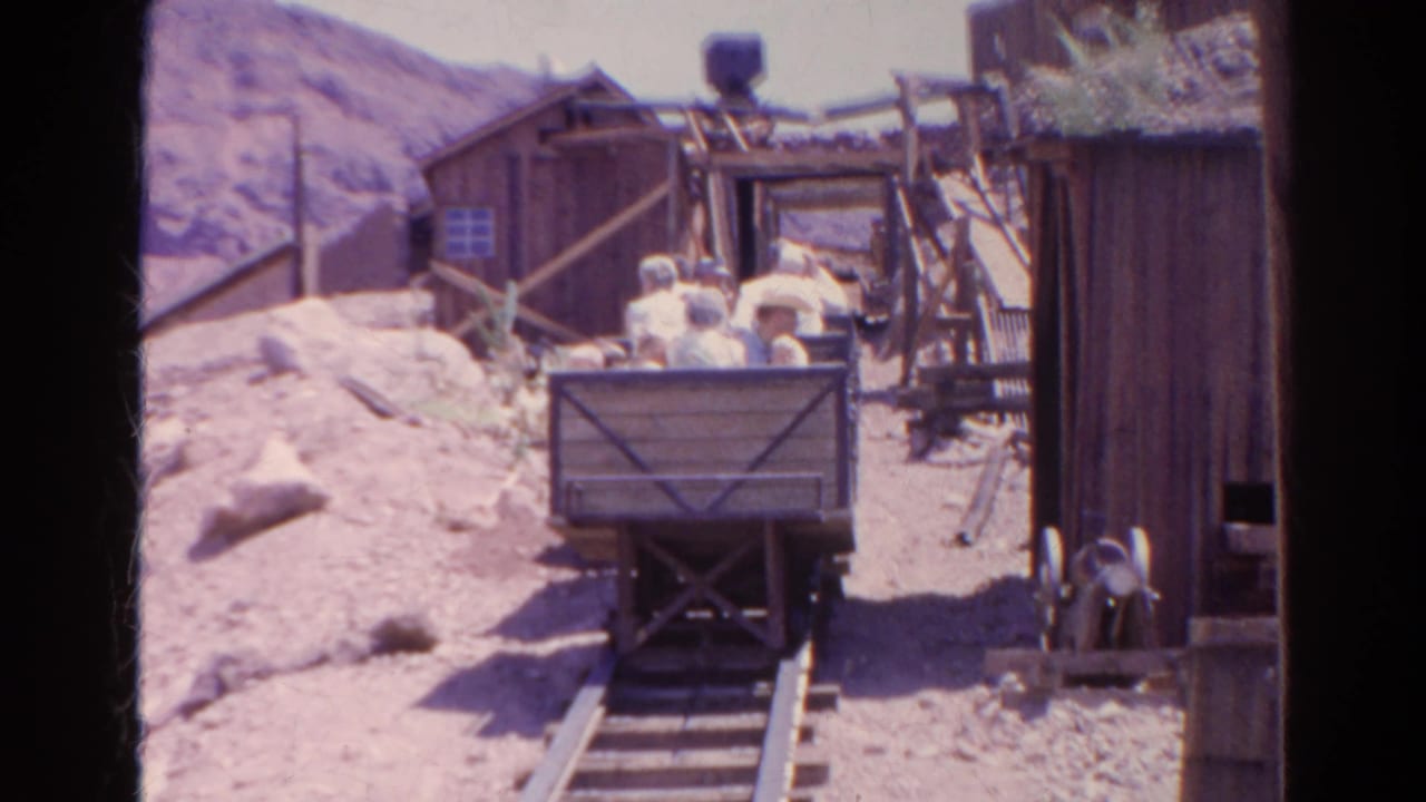 1966 8mm frame from Silver Lake, California, showing men in a wooden mine cart on rails near rustic mining structures and dry hills.