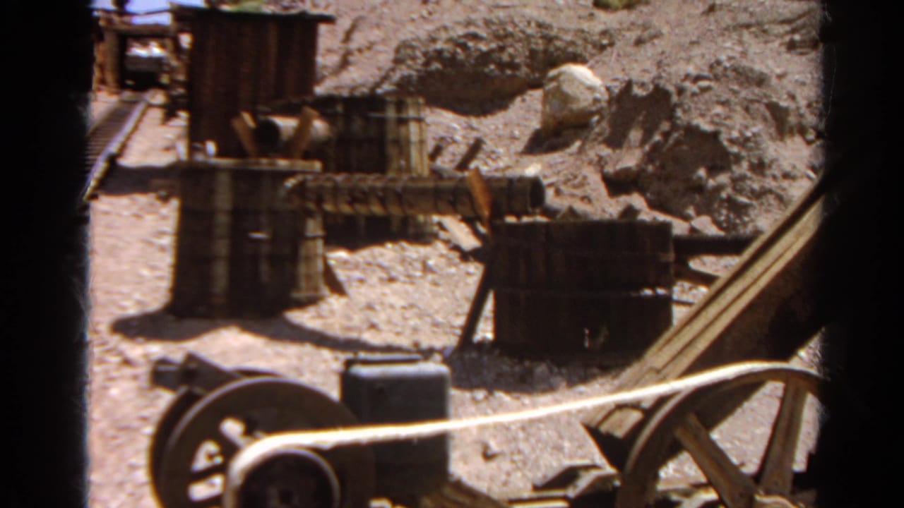 Rocky worksite with barrels, wooden structures, and a large wheel in sunlight.