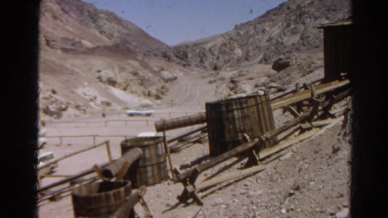 1966 Silver Lake view across a dirt road and rocky hills, with pipes, barrels, and wooden industrial equipment in the foreground.