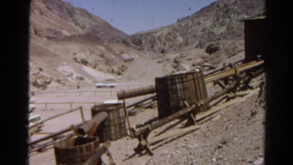 1966 Silver Lake view across a dirt road and rocky hills, with pipes, barrels, and wooden industrial equipment in the foreground.