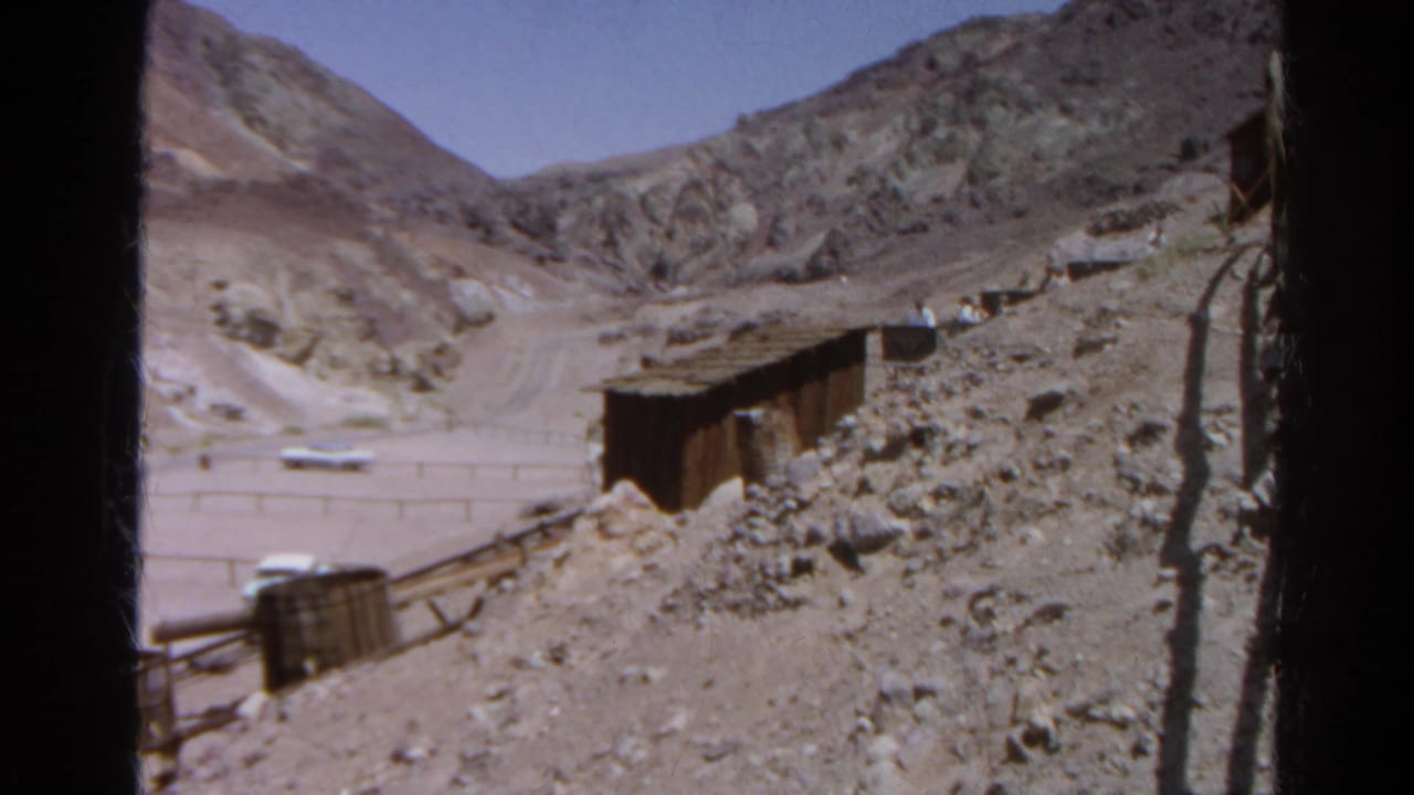 Rocky mining hillside with a small shed, rails, fence, and distant white car.