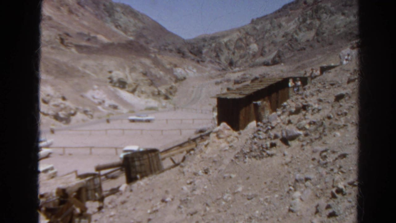 1966 8mm view of a sunlit California desert mining area with a dirt road, weathered wooden structure, and rocky hillside.