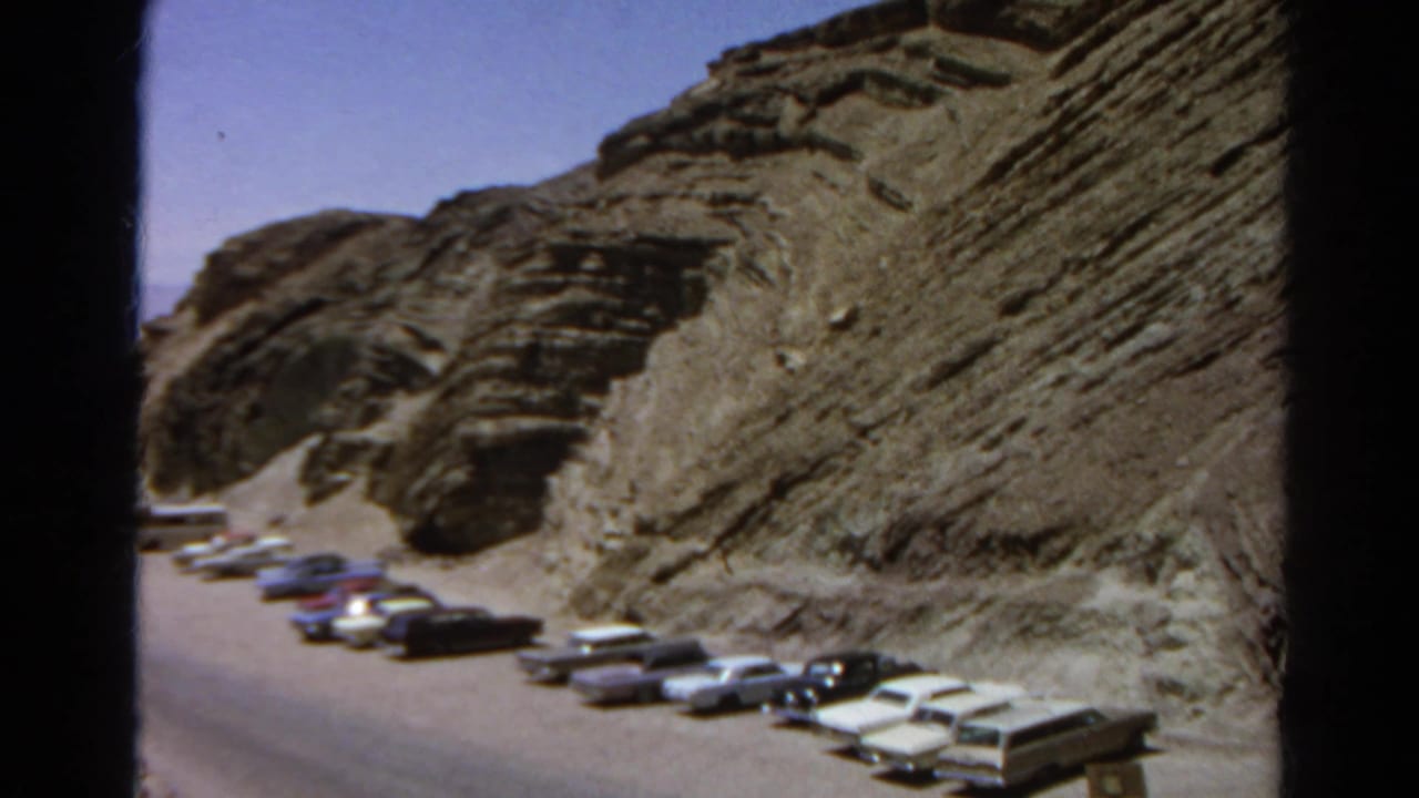 Rows of parked cars along a paved road below a steep rocky hillside.