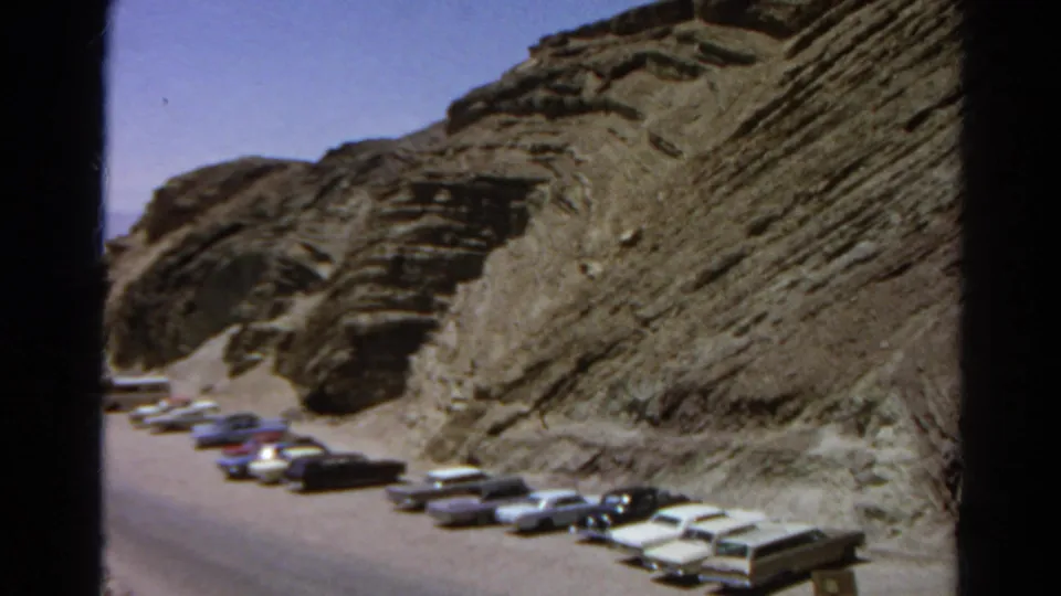 1966 California desert roadside scene with mid-century cars parked in rows beneath a layered rocky slope.