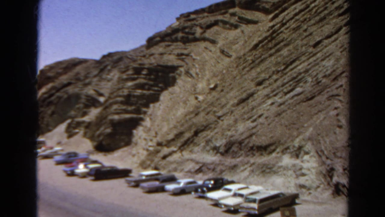 Soft 8mm frame showing a line of 1960s cars near a rugged California desert hillside and roadside turnout.