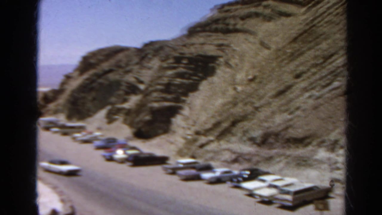 1966 8mm roadside view of classic cars parked near a steep rock face in a California desert setting.