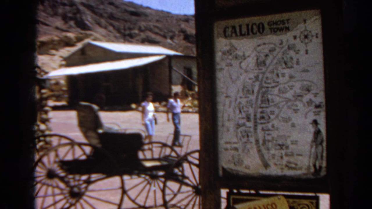 1966 8mm view of Calico Ghost Town with a map sign, carriage display, walking visitors, wooden buildings, and rocky hillside.