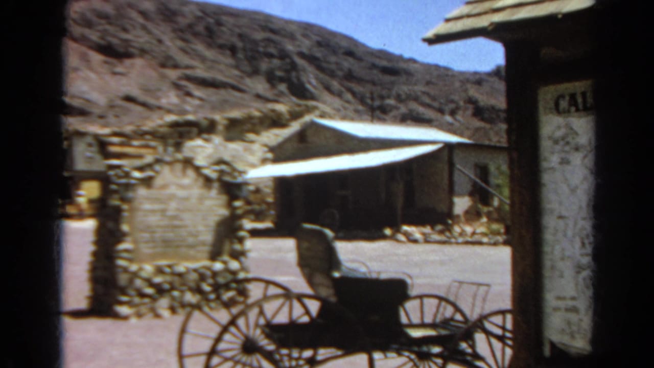1966 8mm frame showing a carriage display, partial Calico Ghost Town sign, stonework, wooden building, and desert hillside.