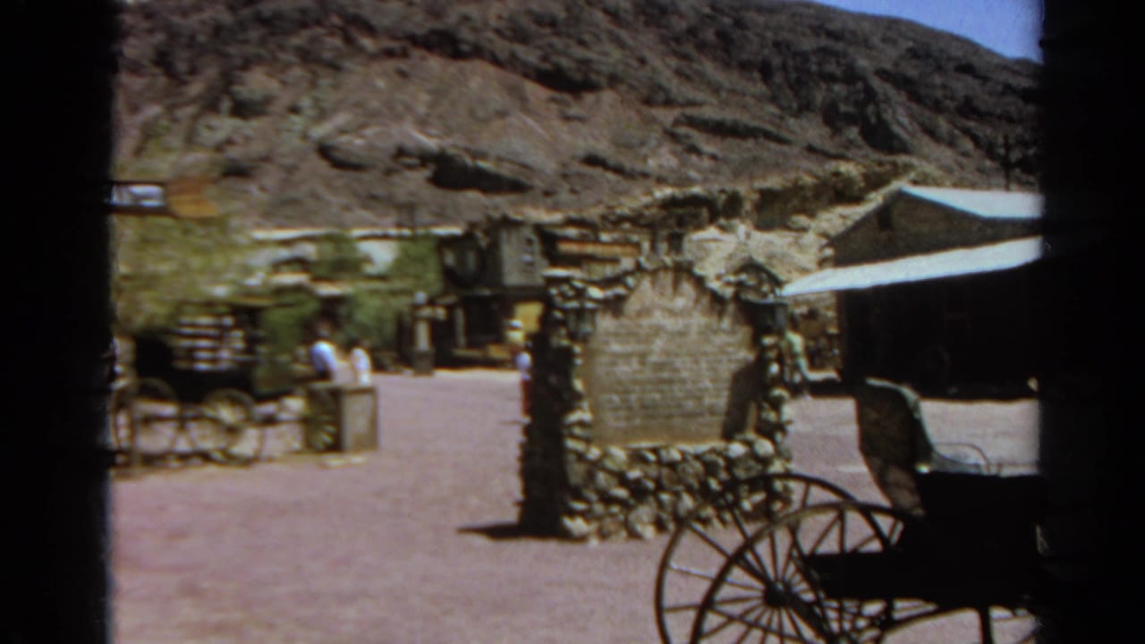 Wide 1966 home-movie view of Calico Ghost Town with visitors near a stone gateway, a carriage display, wooden storefronts, and desert hills.