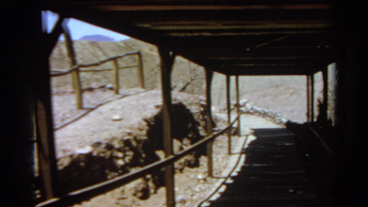 Visitor-perspective 8mm frame from a covered wooden walkway, looking past railings toward a dusty path and rocky hillside in the Silver Lake setting.