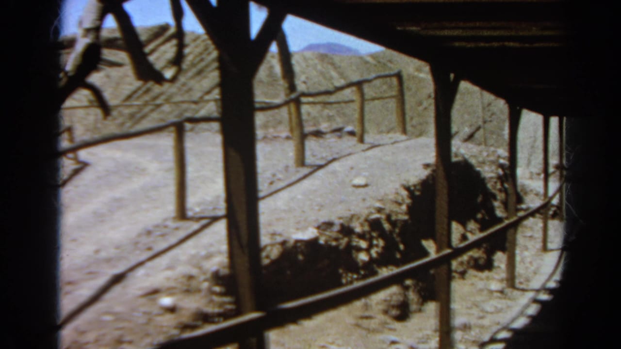 Handheld 8mm view through dark wooden posts and railings toward a dusty path and rocky embankment at the rustic Silver Lake site.