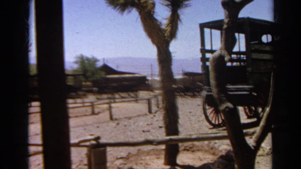 1966 8mm frame from Silver Lake, California, looking through wooden posts toward wagons, rail fencing, a Joshua tree, and arid hills.