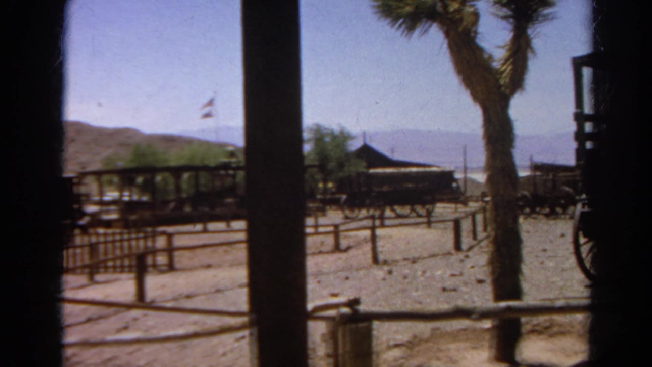 Fence rails, wagons, a flag, trees, and hills seen through dark wooden posts.