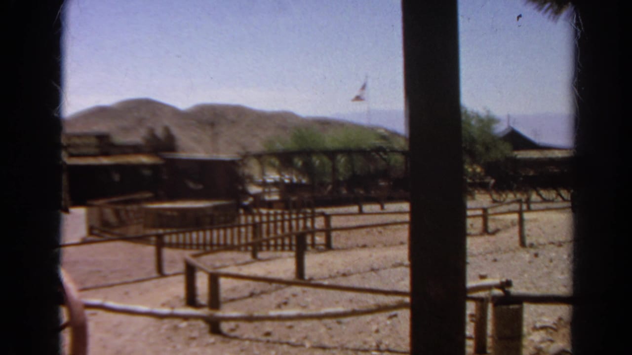 1966 8mm view of a Western-themed yard in Silver Lake, with rail fencing, wagons, foreground posts, and arid hills in the background.