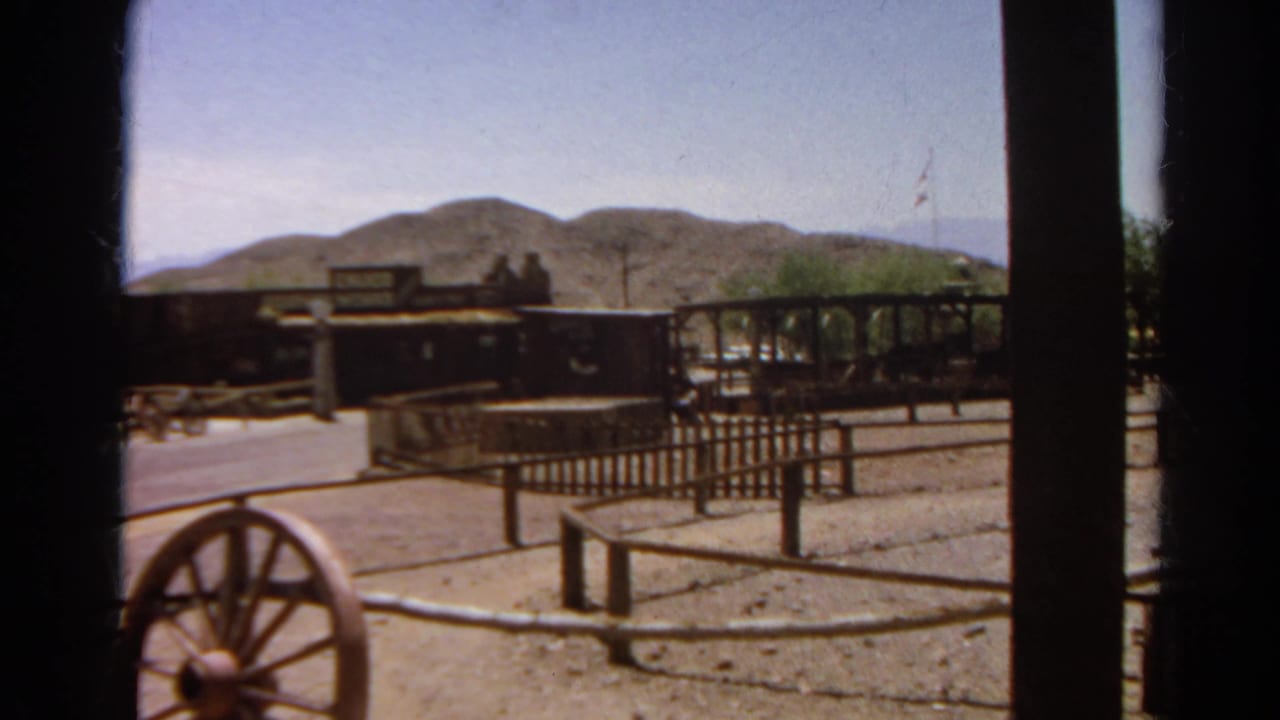 Final 1966 Silver Lake frame with a wagon wheel near the camera, rail fencing, Western-style wagons, trees, and desert hills beyond.