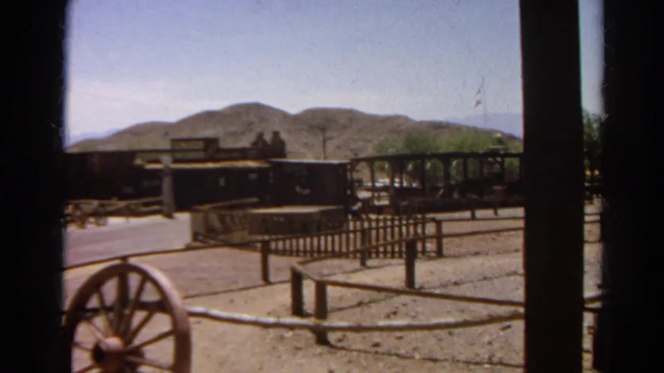 Final 1966 Silver Lake frame with a wagon wheel near the camera, rail fencing, Western-style wagons, trees, and desert hills beyond.