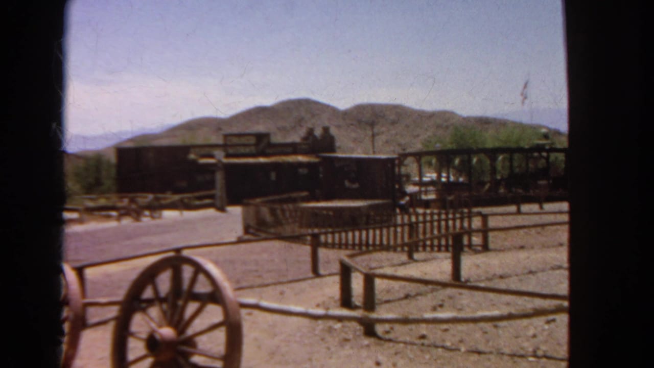1966 8mm view of a California Old West-style street with wagon wheels in the foreground, wooden buildings, fencing, and arid hills.