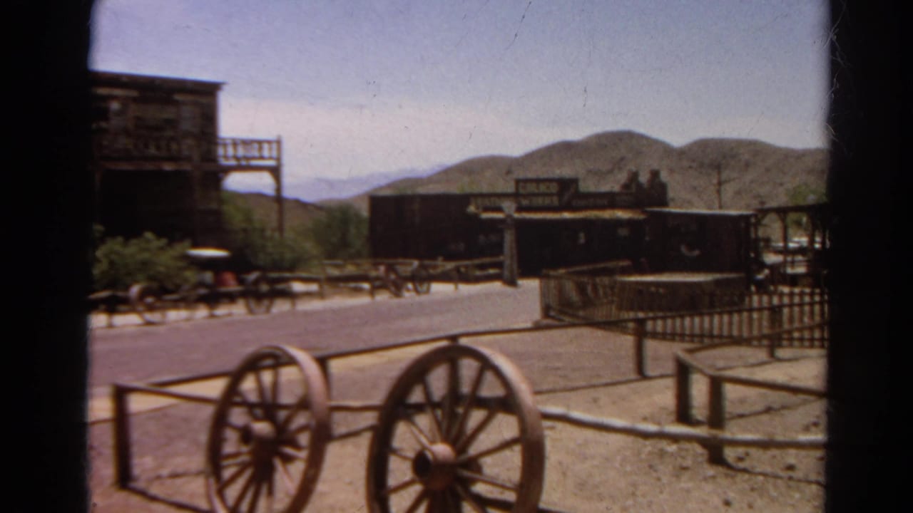 Frontier-style buildings, wagon wheels, rail fence, and distant hills along a quiet street.