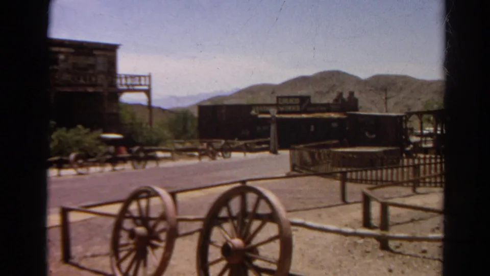 Visitor-shot 1966 8mm frame showing Old West-style storefronts, wagon wheels, fencing, a quiet street, and hills in California.