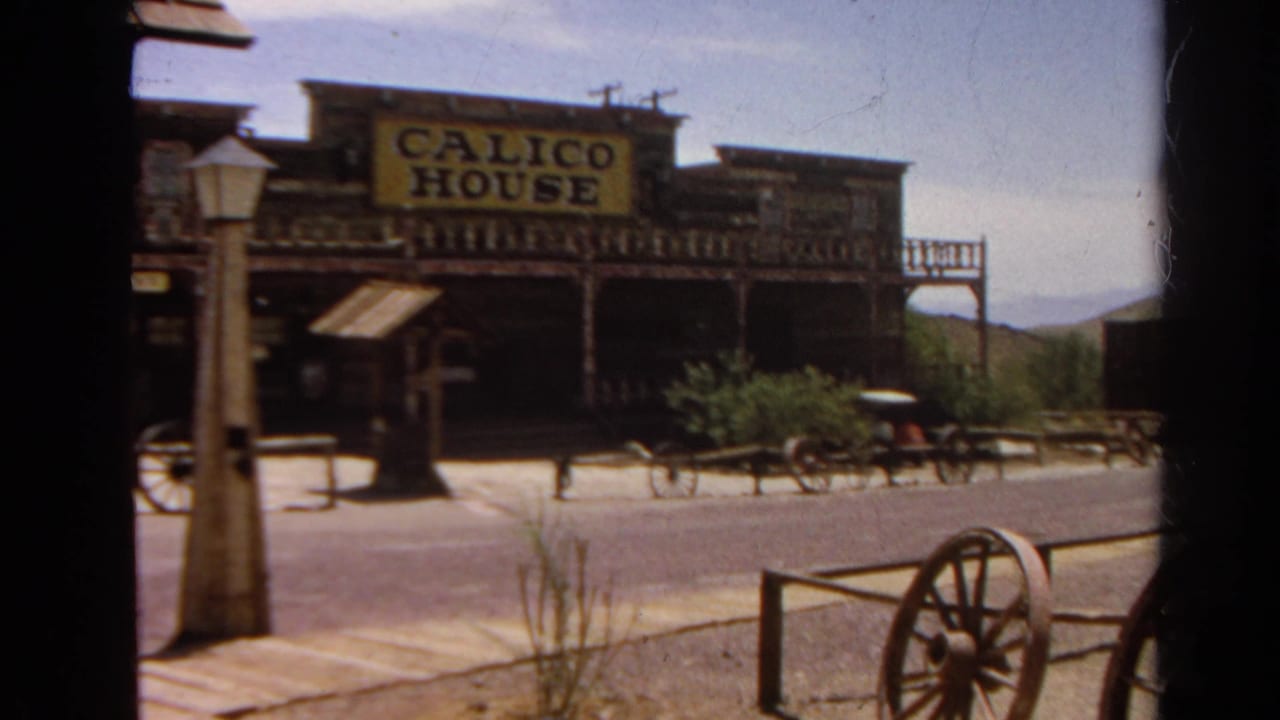 1966 8mm view of the Calico House frontage with sign, wooden posts, a small wagon, street, and hills in the background.
