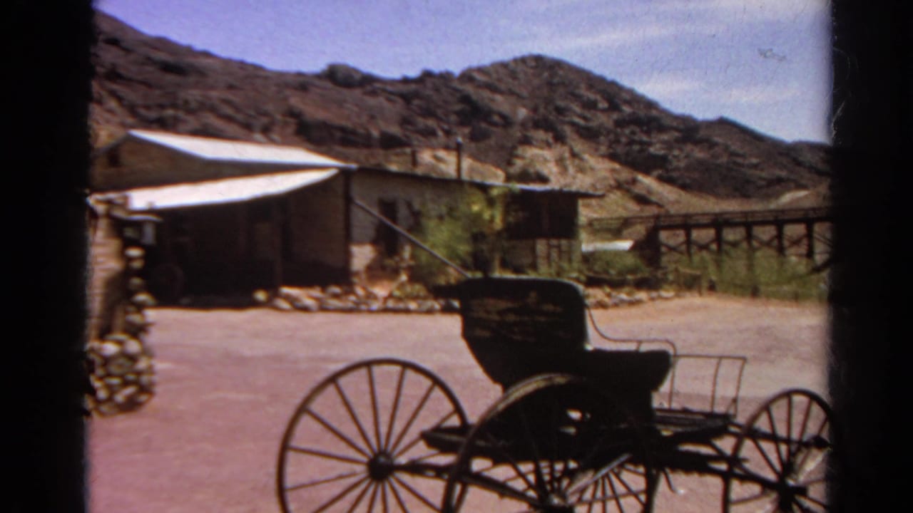1966 8mm view of a wooden wagon, low stone wall, rough structures, and dry hills in a California ghost town setting.
