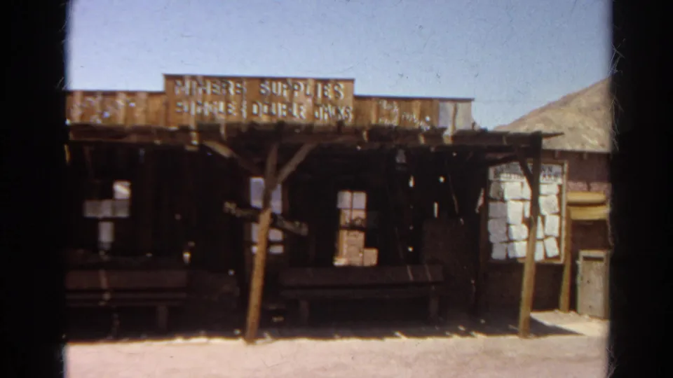 Handheld 8mm frame of the Miners Supplies building, showing rustic wood siding, porch supports, and patched window openings.