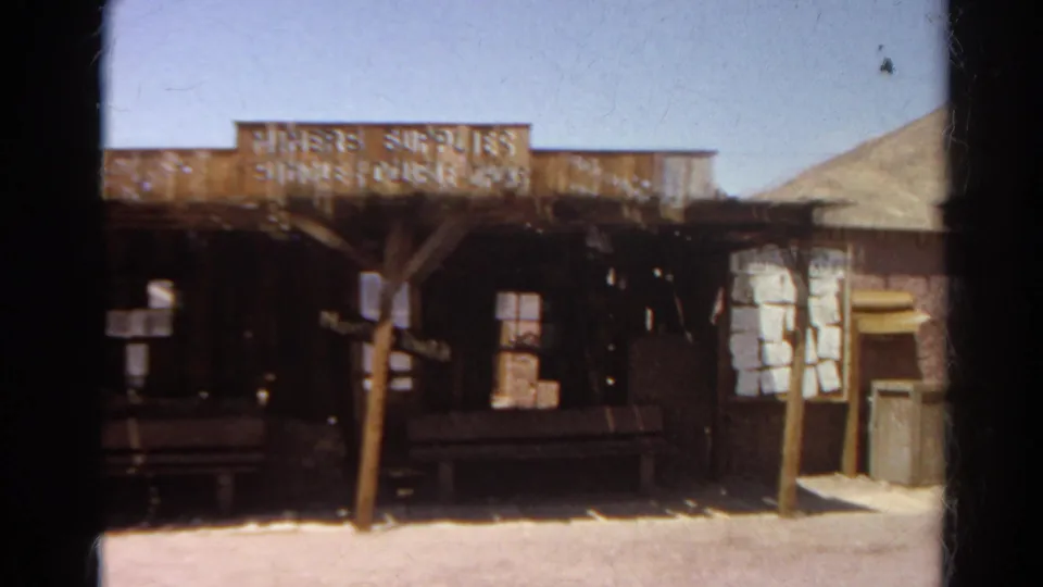 Final 1966 8mm frame of the preserved mining-town storefront, with the sign, porch, windows, desert ground, and pale hillside visible.