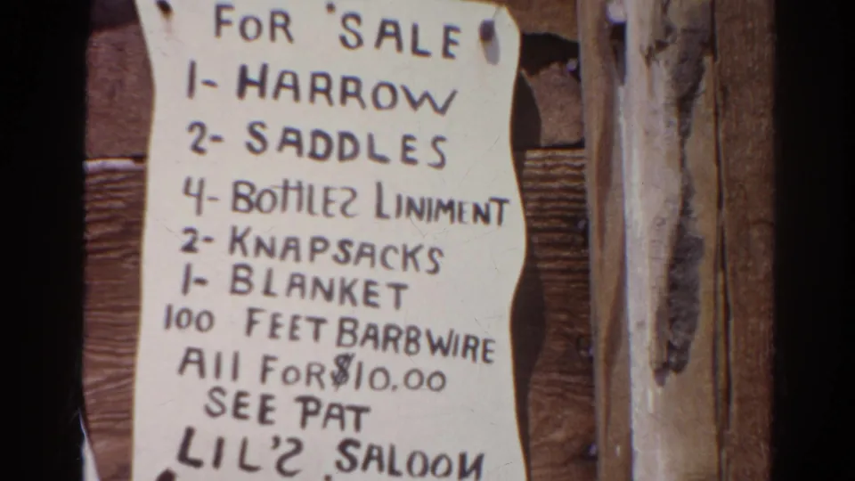 1966 Silver Lake 8mm frame showing a handwritten sale notice for saddles, knapsacks, a blanket, and barbed wire on rough boards.