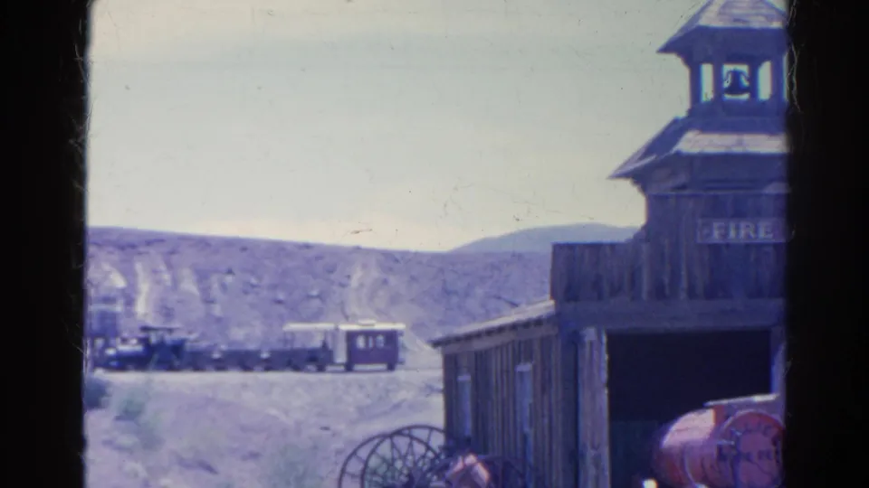 1966 8mm frame from Silver Lake, California showing a Western-style fire hall facade with bell tower, wagon wheel, distant train, and desert hills.