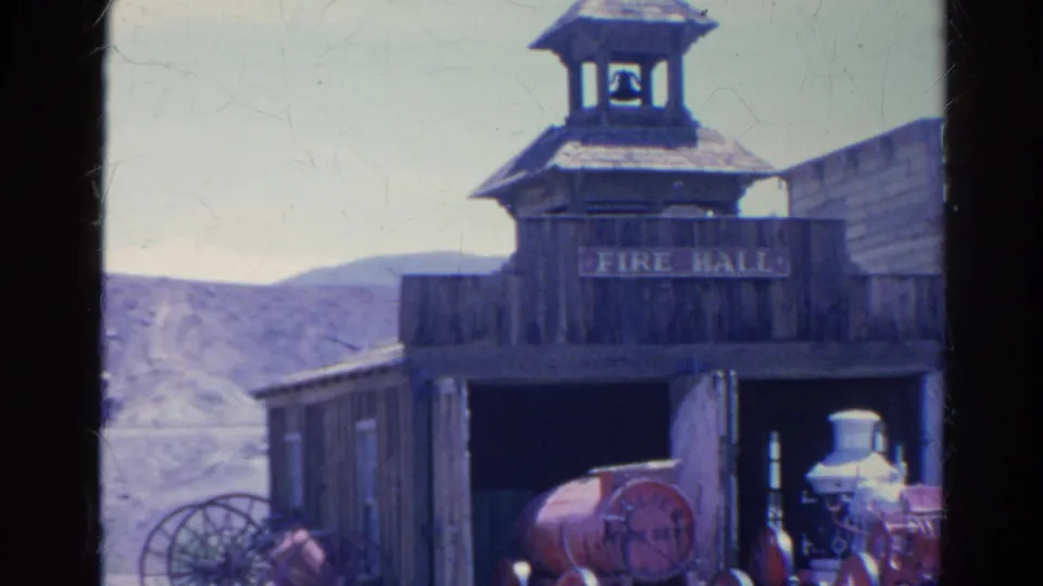 Closer 1966 8mm view of the Silver Lake fire hall facade with FIRE HALL sign, bell tower, wagon wheel, open bay, and red firefighting display.